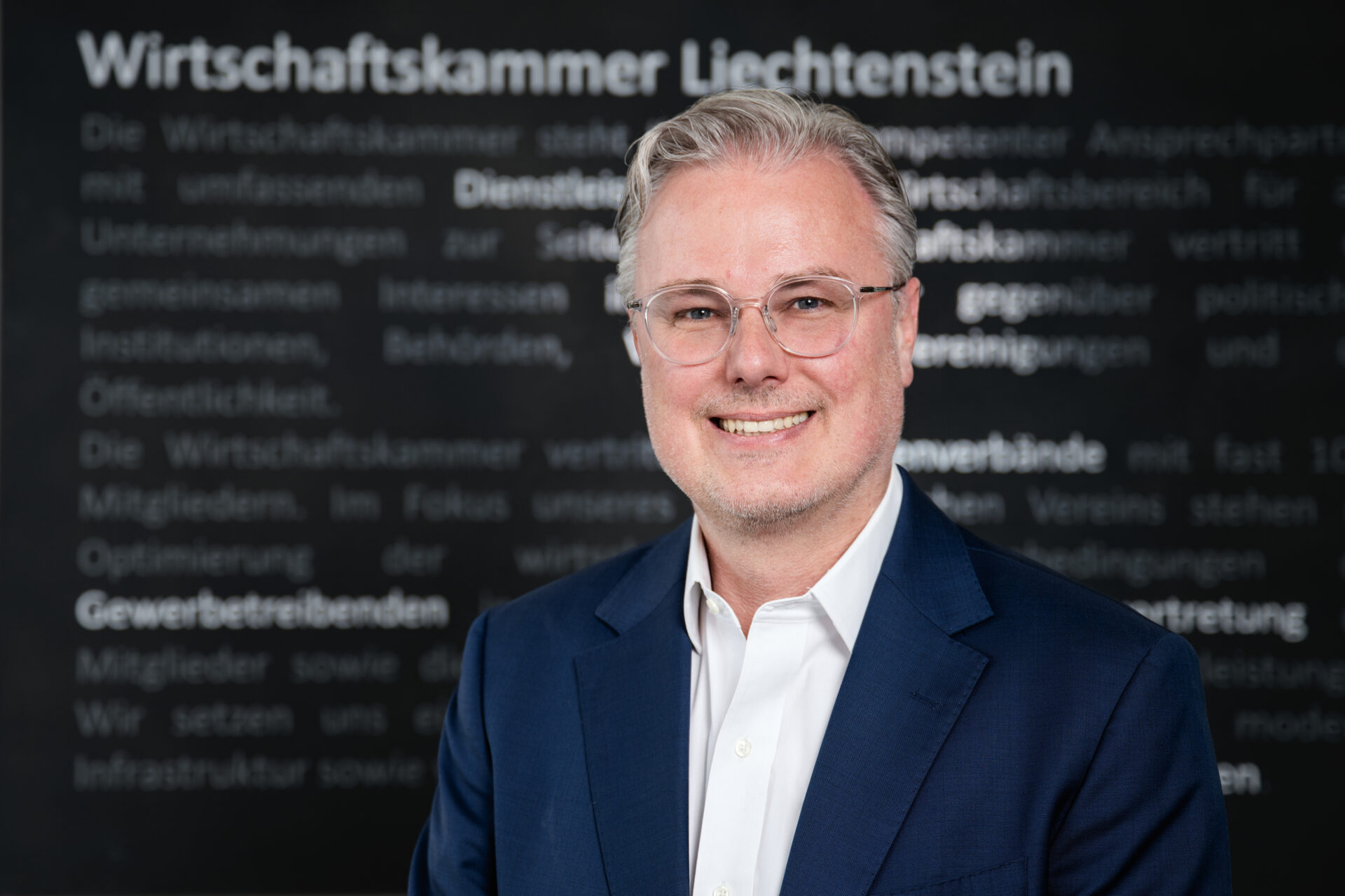 Lächelnder Geschäftsmann in Anzug vor Hintergrund der Wirtschaftskammer Liechtenstein.