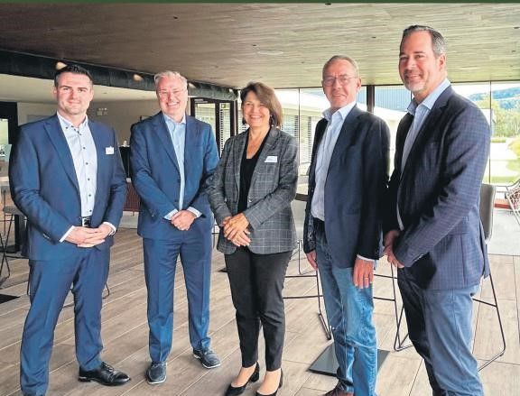 Fünf Fachleute in Anzügen posieren lächelnd bei einem Networking-Event in modernem Raum.