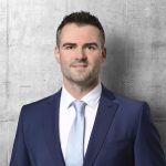 Professioneller Mann in navy Anzug vor einer modernen Betonwand, strahlt Vertrauen und Professionalität aus.